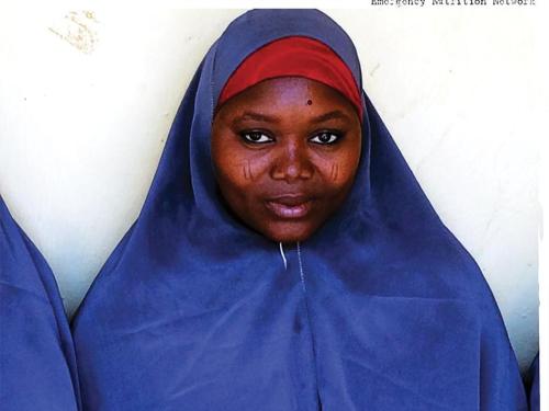 Image shows a woman in a hijab standing against a wall.