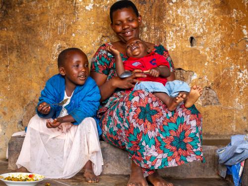 Mother and her two children smiling while the older child eats with a spoon.