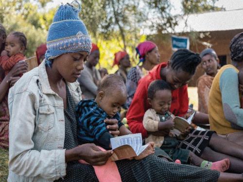 To the fore of a group of people, two women sitting on the grass with infants on their laps looking at a small record book.