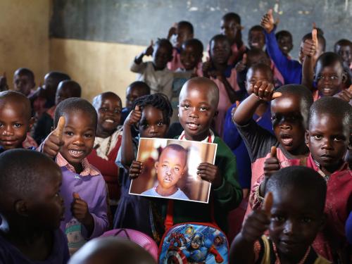 A school child holds up a photo of himself before having had cleft survey, he is surrounded by a group of his peers. 