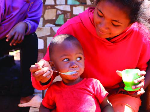 Female wearing a red jumper feeding an infant porridge with a spoon