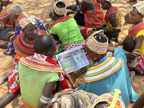 Group of people sitting on the ground, wearing traditional multi-coloured beaded necklaces and headpieces, looking at an A4 sized poster.