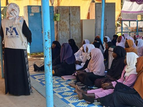A group of women with their young children sitting in a row while a women stands and speaks to them