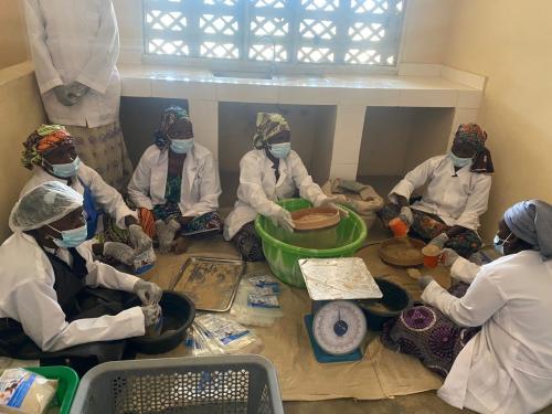 Six women dressed in simple PPE (face masks, doctors coats, gloves etc) sit on a floor siving, measuring up and filling plastic bags with a flour mixture.