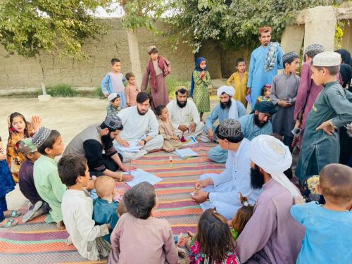 A group of men and children sit and stand outside on a floor mat while looking on to one man who is taking notes. A MUAC tape can also be seen.