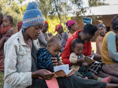 To the fore of a group of people, two women sitting on the grass with infants on their laps looking at a small record book.