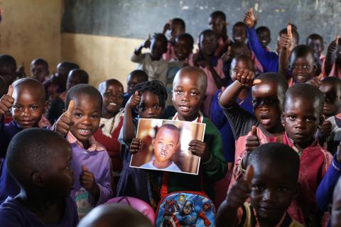 A school child holds up a photo of himself before having had cleft survey, he is surrounded by a group of his peers. 