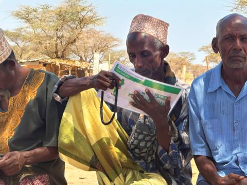 A community elder looks at an A4 sized poster, two other community elders are sat either side of him. 